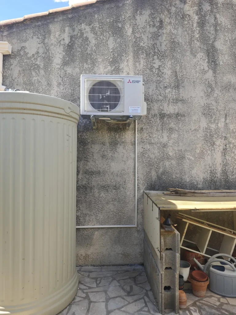 Unité extérieure de clim fixée en façade de cour, en retrait derrière une cuve et des jardinières