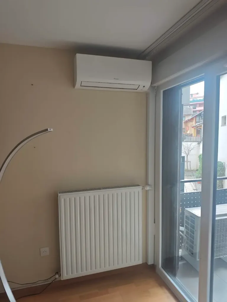 Unité intérieure de climatisation réversible posée au-dessus d’un ancien radiateur, près d’une baie vitrée