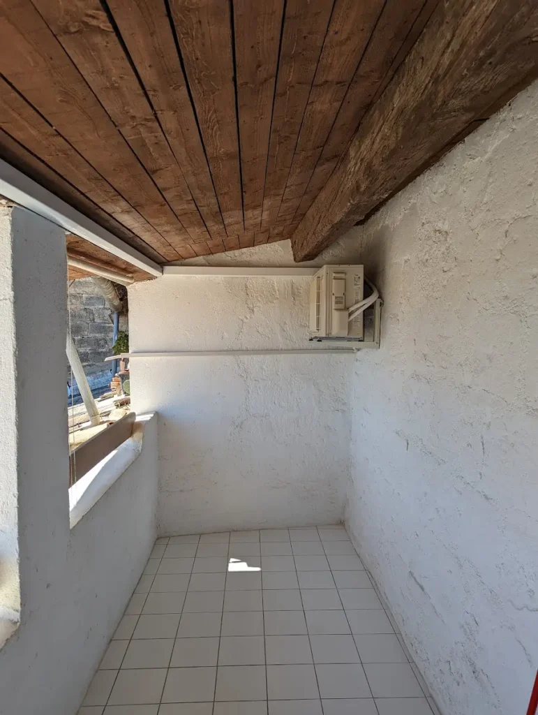 Loggia carrelée sous plafond en bois, avec l’unité extérieure de climatisation posée au fond